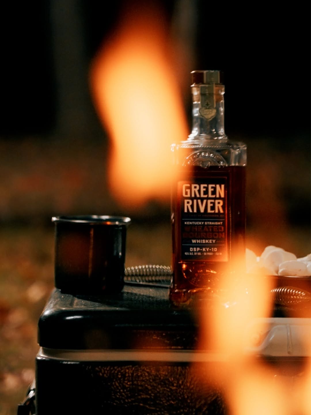 A bottle of Kentucky Straight Wheated Bourbon next to a mug at a campfire.