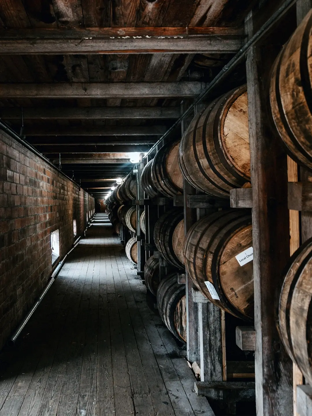 Long wooden rickhouse corridor lined with aging whiskey barrels stacked on racks in a dimly lit warehouse.