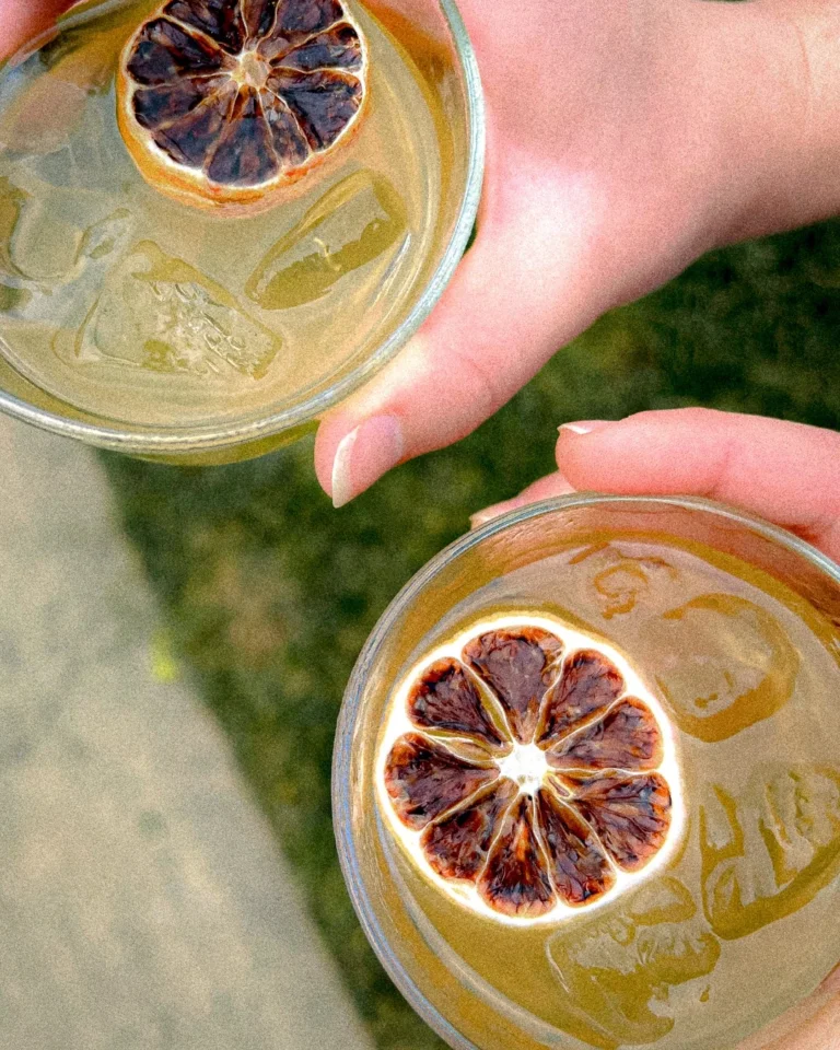 Two hands holding citrus-garnished whiskey cocktails over ice in clear glasses outdoors
