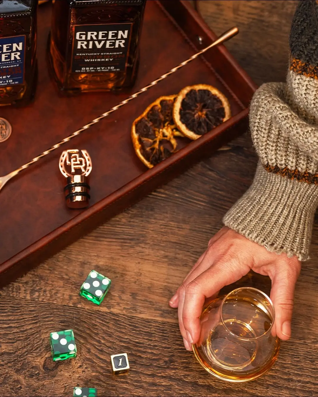 Hand holding a glass of whiskey beside Green River bourbon bottles, dice, and cocktail tools on a wooden table.