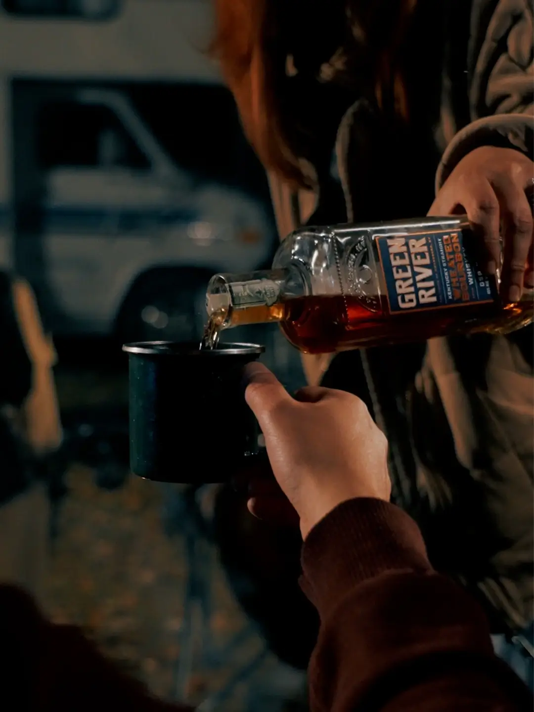 Green River Single Barrel Bourbon being poured into a camping mug beside a campfire during an outdoor evening gathering.
