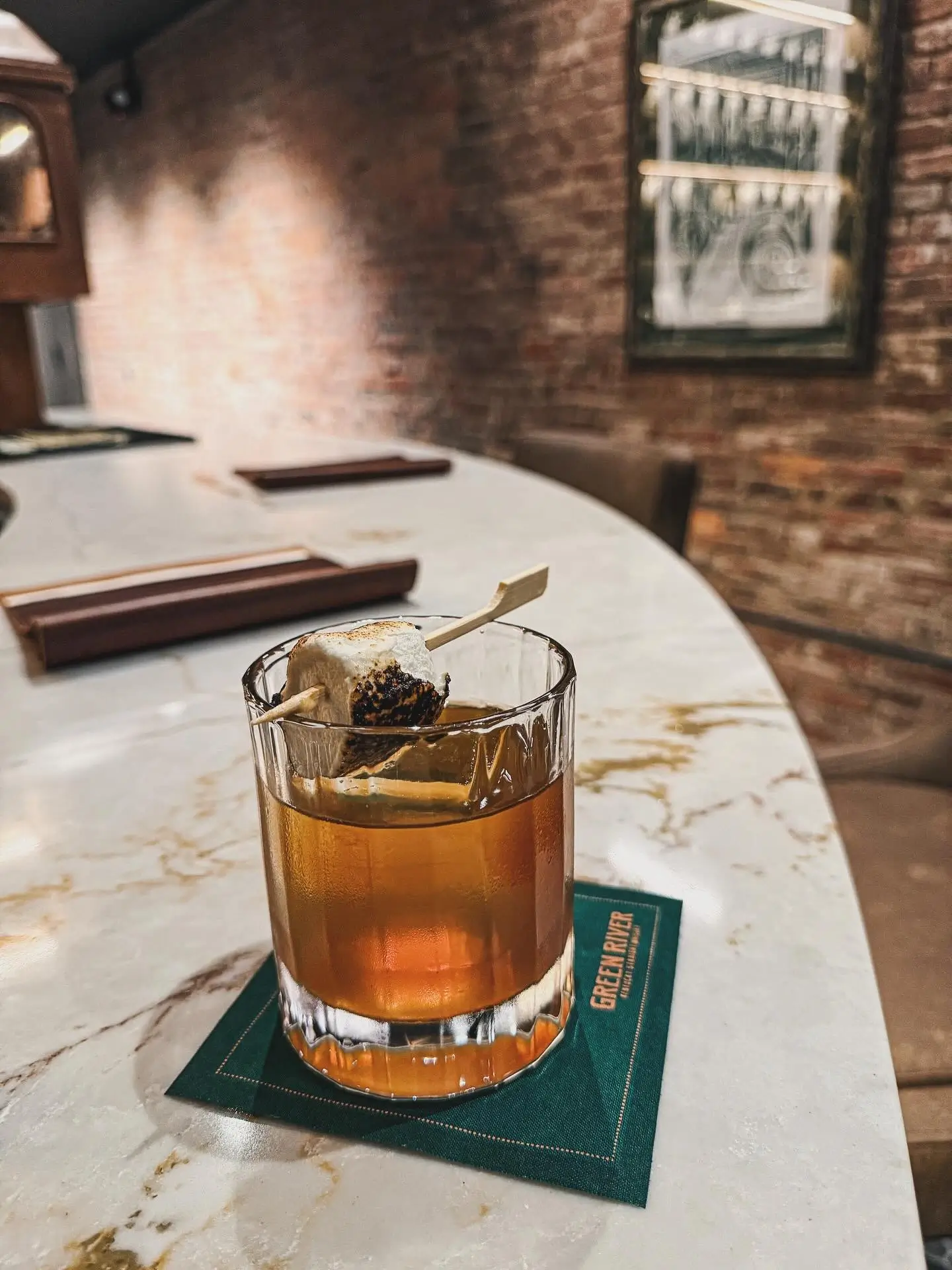 Whiskey cocktail with a toasted marshmallow garnish on a Green River coaster atop a marble bar counter.