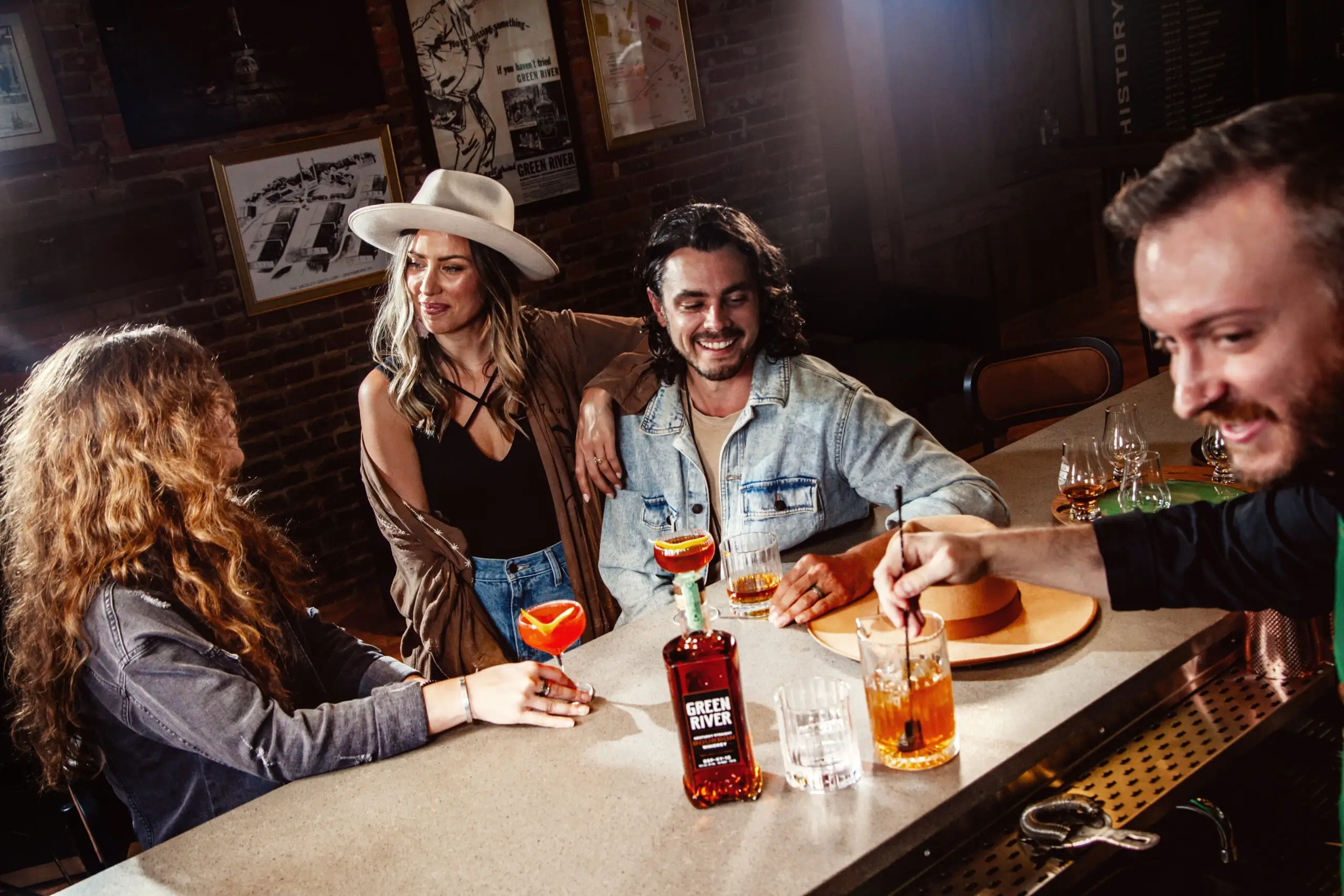 Group of four people toasting with cocktails at a bar counter with Green River whiskey bottle visible.