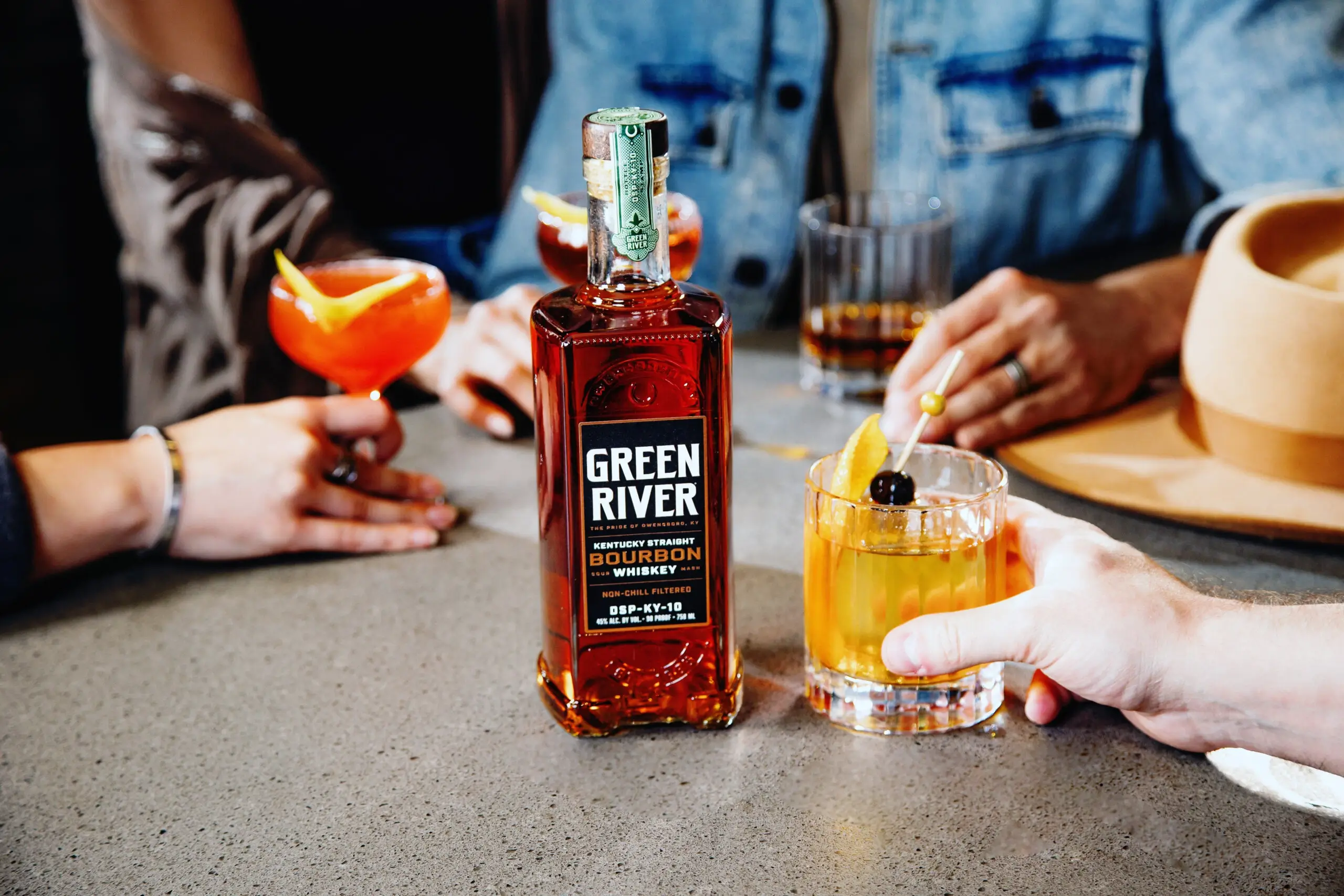 Green River whiskey bottle with cocktail and orange garnish on concrete counter at tasting room gathering.