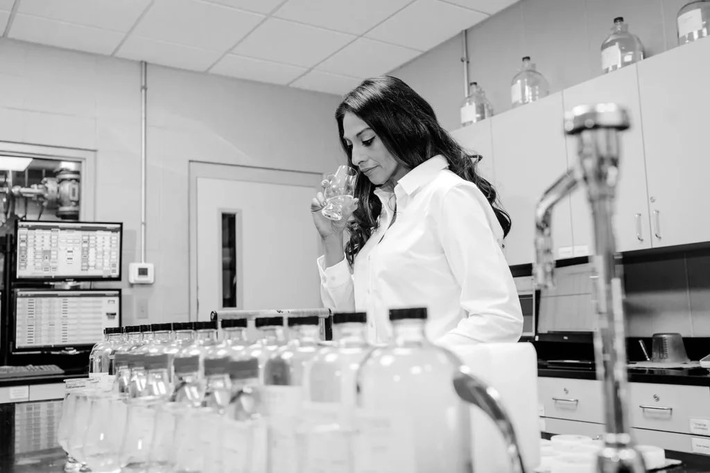 Woman in white lab coat examining glass sample in modern laboratory with workbench and data displays.