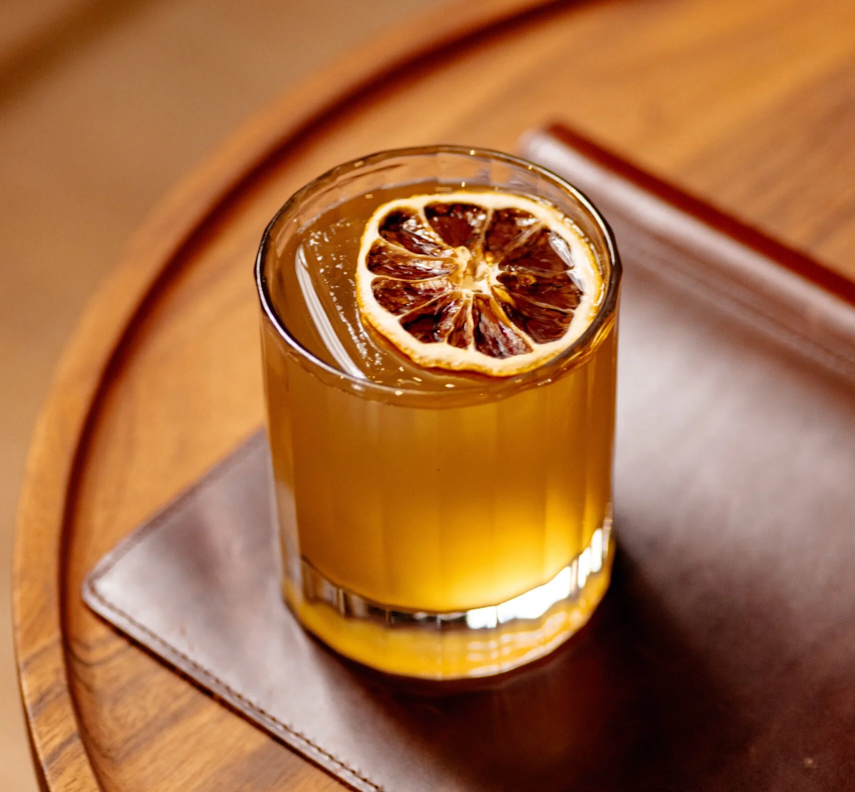 Gold-colored cocktail in rocks glass topped with dried blood orange slice on wooden serving board.